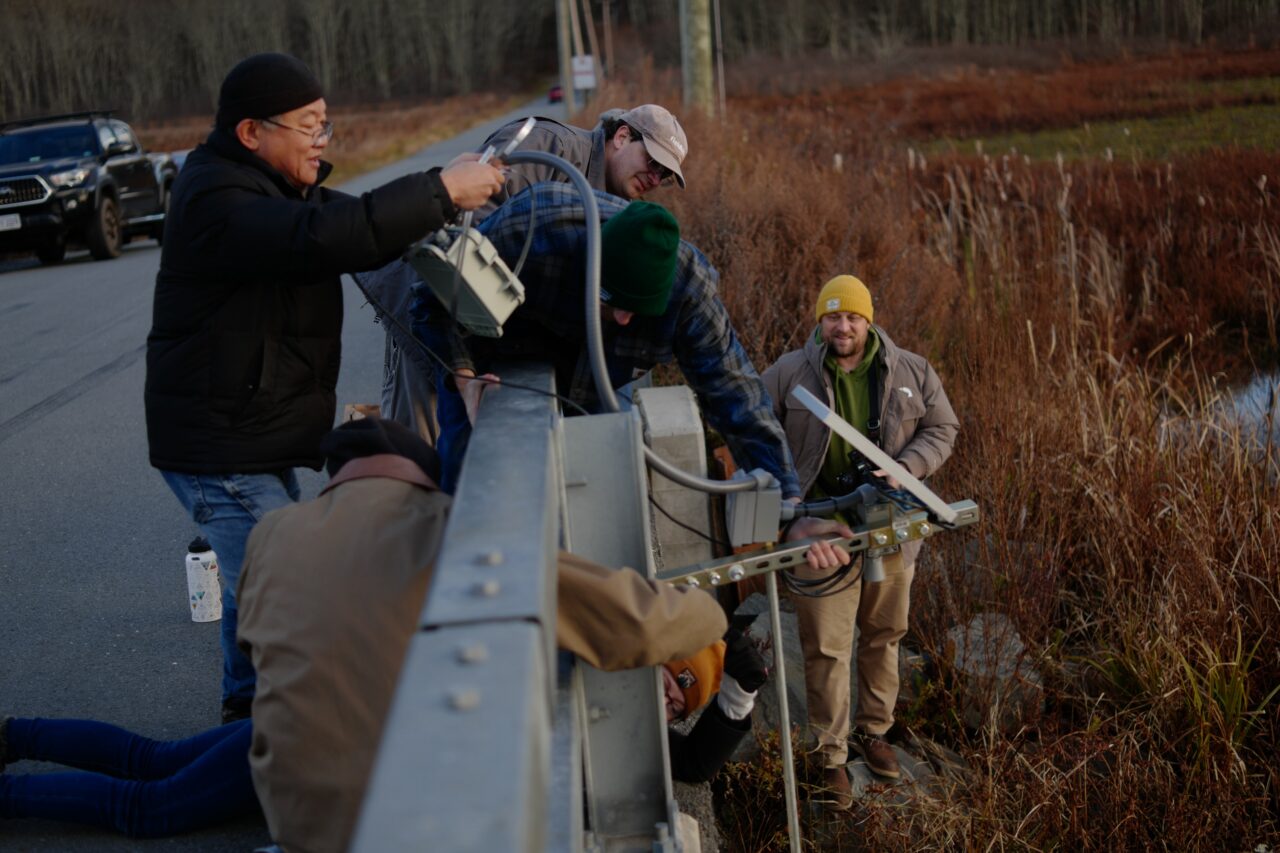 A person holds the FieldKit enclosure while others work to mount the unistrut arm. The cable protector pipe is visible.